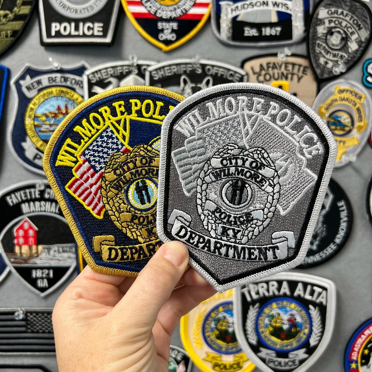 Hand holding full-color and subdued monochrome Wilmore Police Department Kentucky embroidered patches before a wall of agency patches.