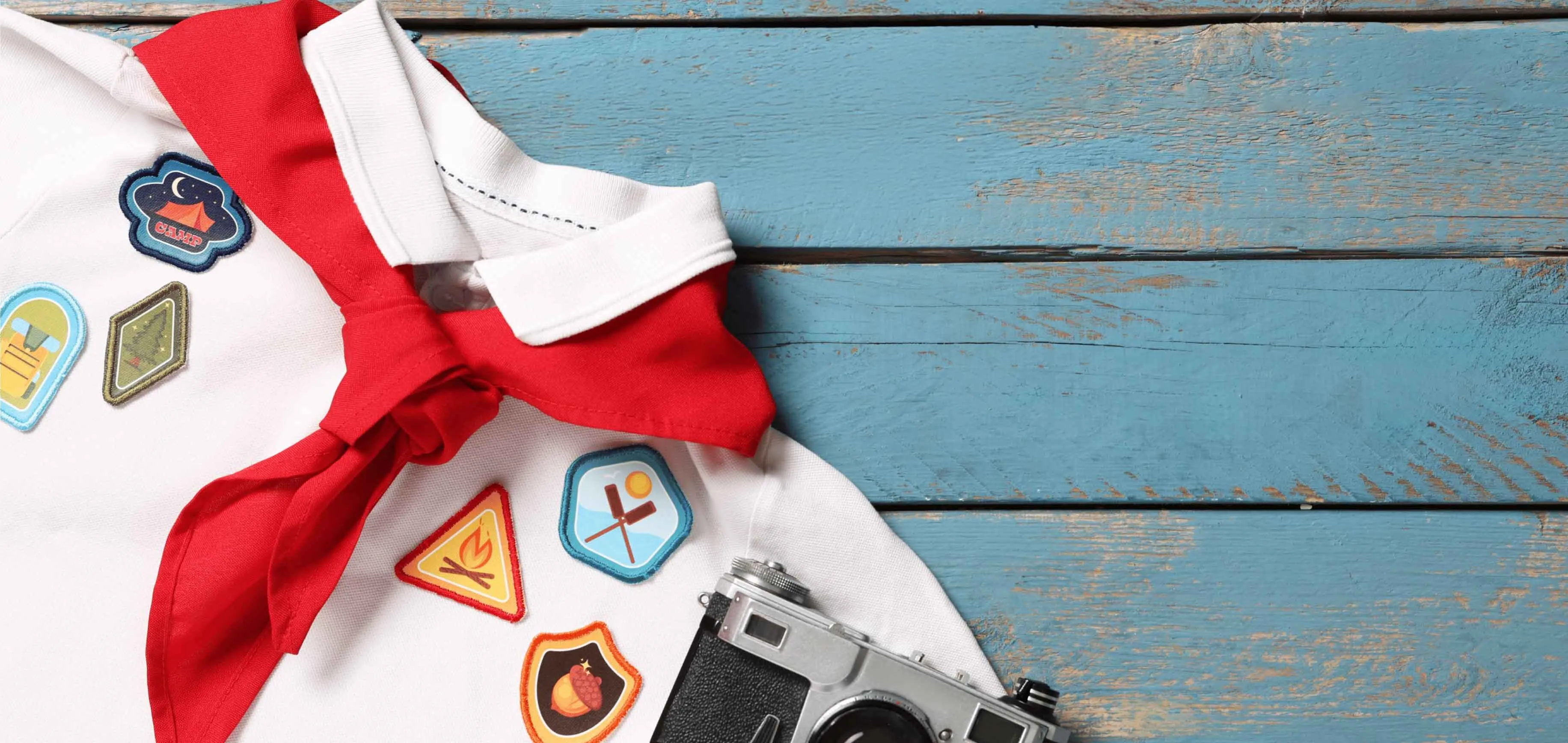 White scout shirt with red neckerchief, colorful merit badges, and camp patches on weathered blue wood beside a vintage camera.