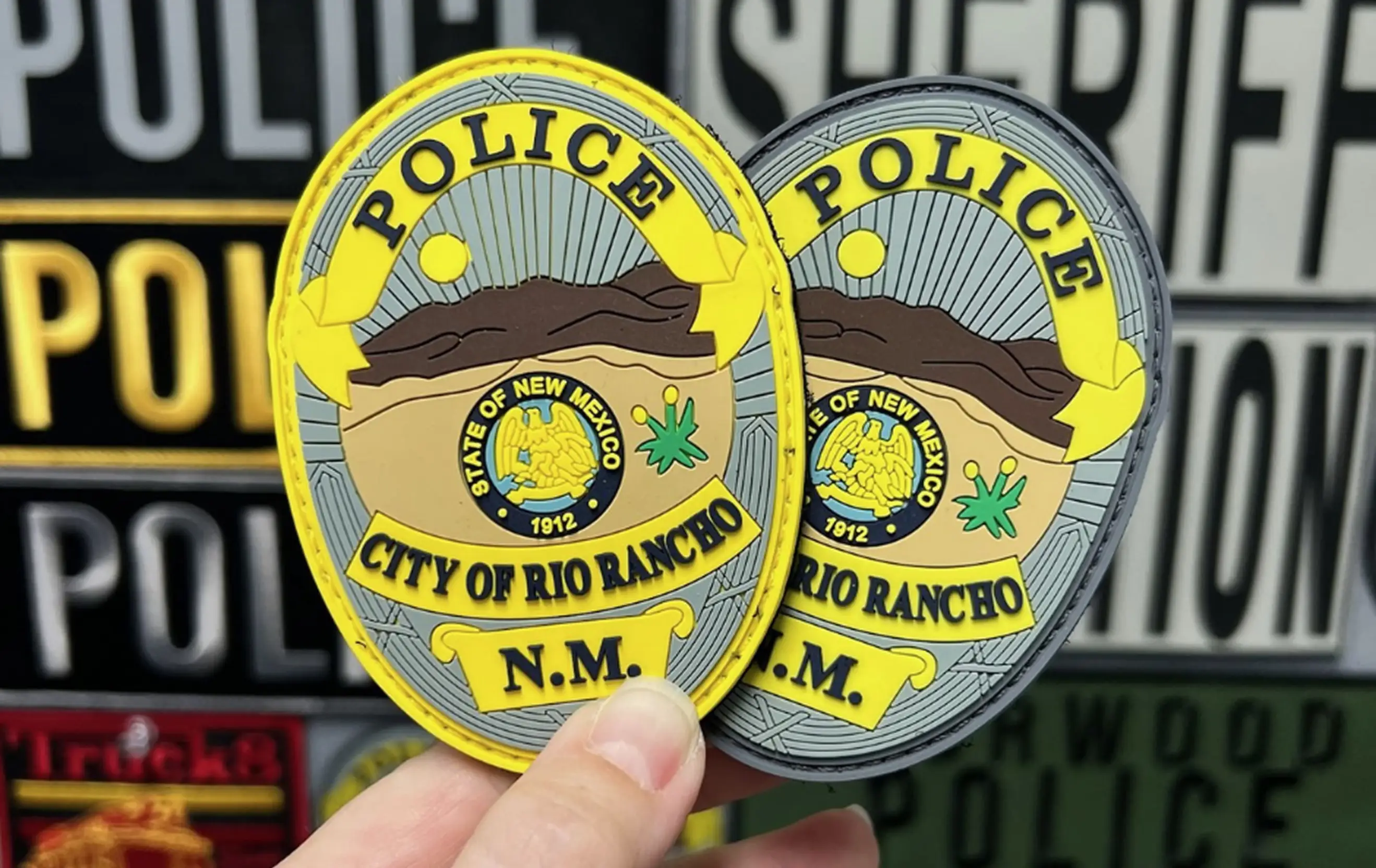 Hands holding yellow- and gray-bordered Rio Rancho New Mexico Police PVC patches with state seal and desert scene.